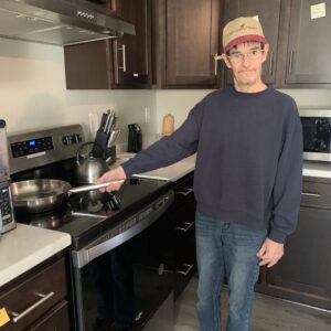 Man in a baseball hat and glasses standing at a stovetop holding a frying pan