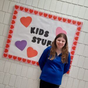 Woman in a blue shirt smiling in front of a sign that says "Kids Stuff"